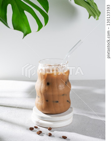 Ice latte coffee in a glass with milk on a light background with coffee beans, monstera leaves Ice latte coffee in a glass with milk on a light background with coffee beans, monstera leaves 130133593