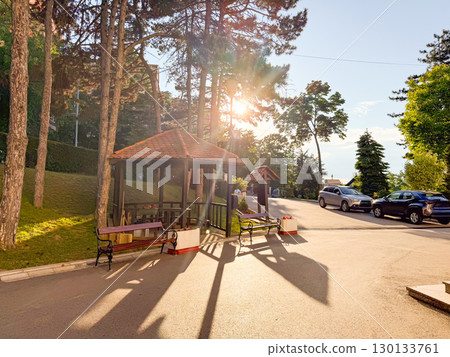 Evening sun shining through pine trees and wooden gazebo with benches near the roadside. Relaxation, summer atmosphere, peaceful rest and connection with nature. 130133761