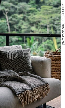 A cozy couch is adorned with a soft blanket and pillow, positioned near large windows that offer a view of lush greenery 130134414