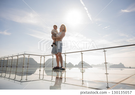 Caucasian woman holds her little son in her arms while standing on a glass bridge in the mountains. Caucasian woman holds her little son in her arms while standing on a glass bridge in the mountains. 130135057