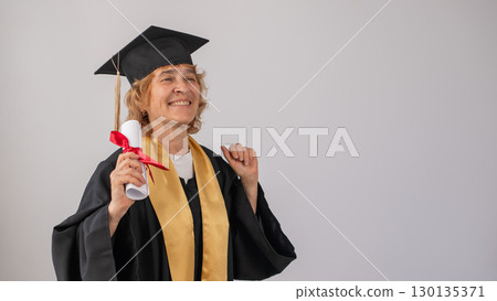 Happy senior woman in graduation gown holding diploma on white background.  130135371