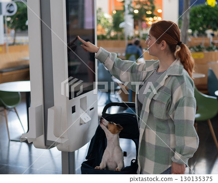Caucasian woman makes a purchase at a self-service counter with a Jack Russell terrier dog in a stroller in a shopping center. Caucasian woman makes a purchase at a self-service counter with a Jack Russell terrier dog in a stroller in a shopping center. 130135375