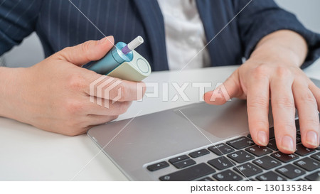 Business woman smoking an electronic cigarette while working on a laptop. Tobacco heating system. Business woman smoking an electronic cigarette while working on a laptop. Tobacco heating system. 130135384