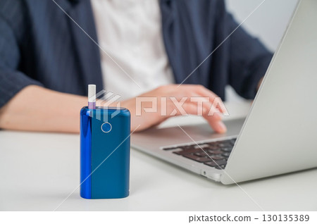 Business woman smoking an electronic cigarette while working on a laptop. Tobacco heating system in the foreground. Business woman smoking an electronic cigarette while working on a laptop. Tobacco heating system in the foreground. 130135389