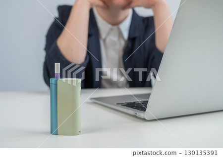 Tired businesswoman smoking an electronic cigarette while working on a laptop. Tobacco heating system. 130135391