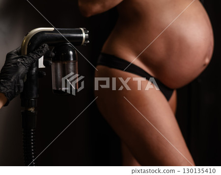 Close-up of the belly of a pregnant woman undergoing an instant tanning procedure. The master applies bronzer with a spray. Close-up of the belly of a pregnant woman undergoing an instant tanning procedure. The master applies bronzer with a spray. 130135410
