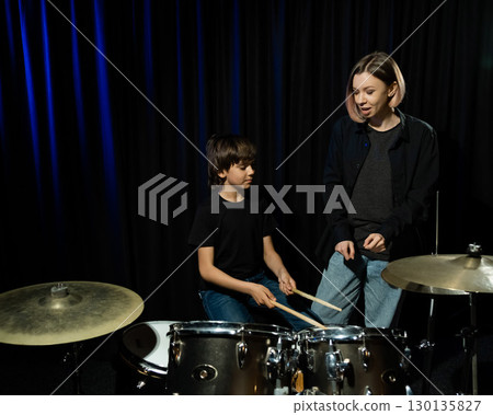 Young caucasian woman teaches a boy to play the drums in the studio on a black background. Music school student Young caucasian woman teaches a boy to play the drums in the studio on a black background. Music school student 130135827