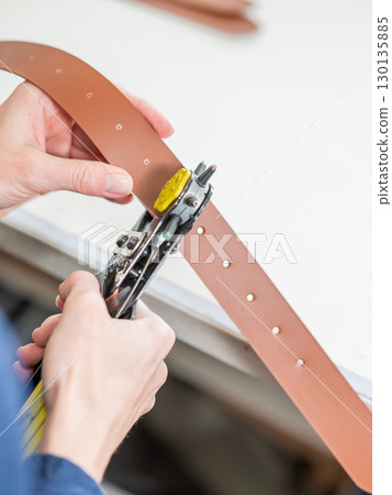 A woman makes holes in a leather belt. Leatherworker's workshop.  130135885