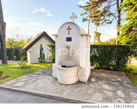 White stone fountain with Christian cross and engraved plaque surrounded by trees and greenery. Tradition, spirituality, religion, cultural heritage and community identity. White stone fountain with Christian cross and engraved plaque surrounded by trees and greenery. Tradition, spirituality, religion, cultural heritage and community identity. 130135958