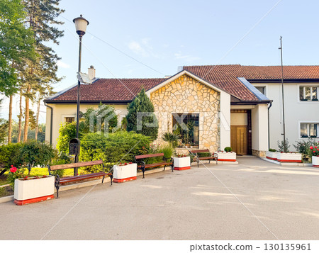 House with stone facade, tiled roof, benches and green garden in residential area on sunny day. Architecture, lifestyle, outdoor recreation and community culture. House with stone facade, tiled roof, benches and green garden in residential area on sunny day. Architecture, lifestyle, outdoor recreation and community culture. 130135961