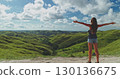 Female tourist standing with open arms, embracing the stunning landscape of rolling green hills and blue sky with clouds in Sumba Island, Indonesia, during a sunny summer day 130136675