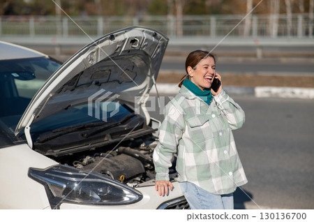 Caucasian woman laughing and calling on phone asking for help standing near car with open hood. Caucasian woman laughing and calling on phone asking for help standing near car with open hood. 130136700