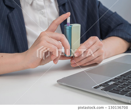 Business woman smoking an electronic cigarette while working on a laptop. Tobacco heating system. Business woman smoking an electronic cigarette while working on a laptop. Tobacco heating system. 130136708