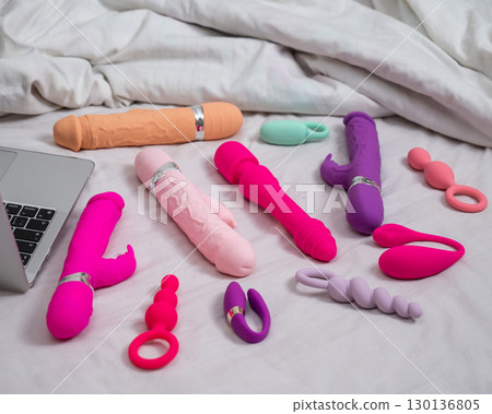 Collection of sex toys on a white sheet. Various vibrators and a laptop on the bed.  130136805