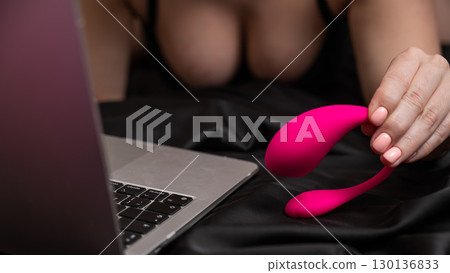 A woman is lying in bed holding a curved vibrator and looking at a laptop.  130136833