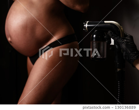Close-up of the belly of a pregnant woman undergoing an instant tanning procedure. The master applies bronzer with a spray. Close-up of the belly of a pregnant woman undergoing an instant tanning procedure. The master applies bronzer with a spray. 130136889