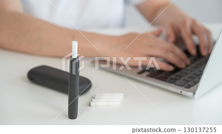 A woman smokes an electronic cigarette while working on a laptop. Tobacco heating system. 130137255