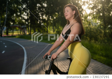 A Caucasian woman in yellow leggings and a crop top rides a bicycle in tights in the park.  130137291