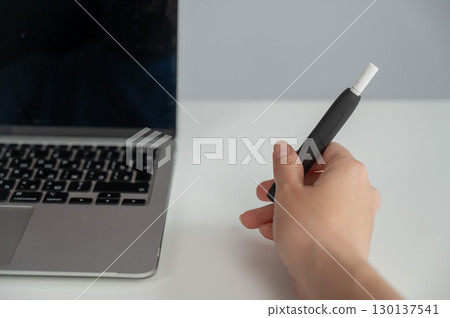 A woman smokes an electronic cigarette while working on a laptop. Tobacco heating system. A woman smokes an electronic cigarette while working on a laptop. Tobacco heating system. 130137541