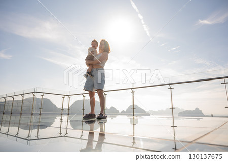Caucasian woman holds her little son in her arms while standing on a glass bridge in the mountains.  130137675