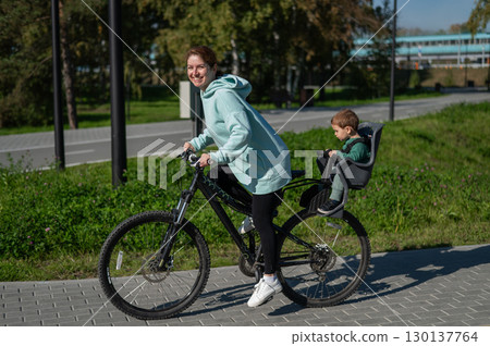 Caucasian woman riding bicycle with toddler son sitting behind her in child seat in park. Caucasian woman riding bicycle with toddler son sitting behind her in child seat in park. 130137764