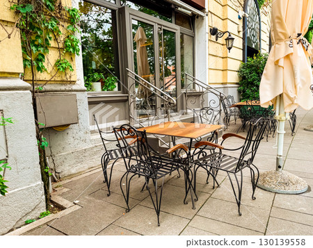 Cafe terrace with wooden tables, umbrellas and decorative chairs near vintage building facade. Outdoor dining, architecture, hospitality, lifestyle and urban culture. 130139558