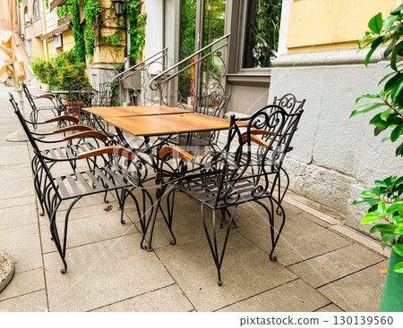 Outdoor cafe terrace with wooden tables and decorative metal chairs near old building. Restaurant, hospitality, lifestyle, urban culture and street dining. Outdoor cafe terrace with wooden tables and decorative metal chairs near old building. Restaurant, hospitality, lifestyle, urban culture and street dining. 130139560