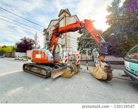 Orange excavator with double bucket parked on residential street under summer sunlight near houses. Engineering, construction, technology, infrastructure repair and urban modernization. 130139587