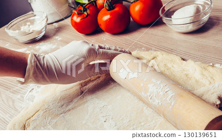 Baker kneading fresh pasta dough with rolling pin surrounded by ripe tomatoes and kitchen bowls on rustic wooden counter Baker kneading fresh pasta dough with rolling pin surrounded by ripe tomatoes and kitchen bowls on rustic wooden counter 130139651