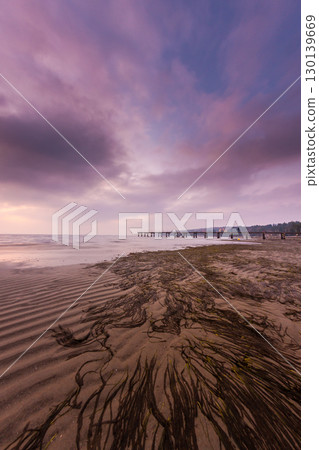Dramatic Purple Sunset Clouds Reflecting Over Tranquil Ocean Beach With Wooden Pier and Seaweed Patterns in Sand 130139669
