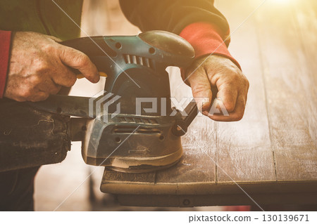 Skilled Carpenter Using Electric Orbital Sander on Wooden Plank in Sunlit Workshop 130139671