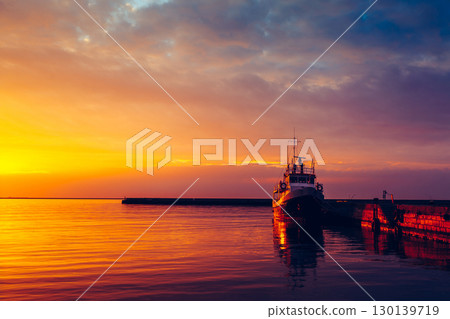 Cargo Vessel Silhouetted Against Brilliant Orange Sunset Reflections on Tranquil Harbor Waters 130139719