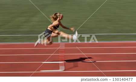 Female athlete sprinting and jumping hurdle with motion blur on stadium track 130140477