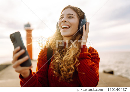 Happy woman listening nice music by the waterfront while wearing headphones by a lighthouse. Happy woman listening nice music by the waterfront while wearing headphones by a lighthouse. 130140487