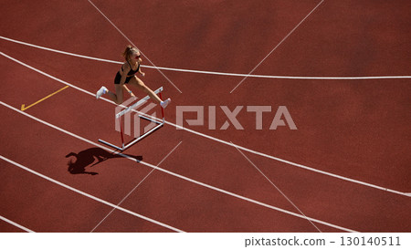 Female track athlete sprinting and clearing hurdle in powerful action motion Female track athlete sprinting and clearing hurdle in powerful action motion 130140511
