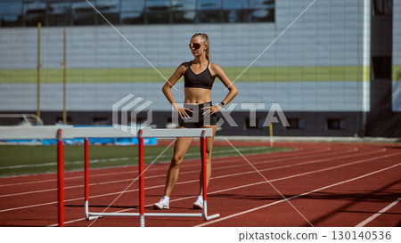 Confident female hurdler standing on track with hurdles Confident female hurdler standing on track with hurdles 130140536
