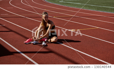 Female runner sitting on track lacing spikes with focus before hurdles training 130140544
