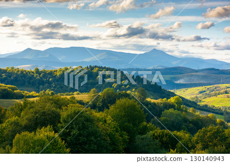 countryside landscape of transcarpathia on september evening. village in the valley and rural fields on the forested hills. carpathian mountain ridge with pikui peak in the distance countryside landscape of transcarpathia on september evening. village in the valley and rural fields on the forested hills. carpathian mountain ridge with pikui peak in the distance 130140943