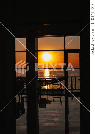 A beautiful sunset over the ocean, viewed from inside a building through the panoramic window. The setting sun casts a golden glow over the sea, seen through a window from inside the restaurant resort 130141129
