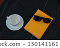 Sunglasses and a yellow book with a cup on a dark, marble table. A cup and saucer, sunglasses, and a yellow book rest on a dark, patterned surface, creating a modern, aesthetic composition. 130141161