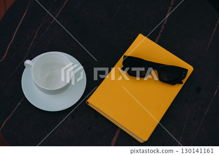 Sunglasses and a yellow book with a cup on a dark, marble table. A cup and saucer, sunglasses, and a yellow book rest on a dark, patterned surface, creating a modern, aesthetic composition. 130141161