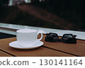 A cup of coffee with sunglasses on a wooden table outdoors. A white teacup and saucer sit on a wooden surface next to sunglasses with a blurred background. 130141164