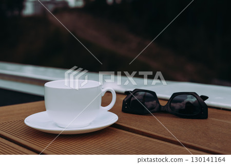 A cup of coffee with sunglasses on a wooden table outdoors. A white teacup and saucer sit on a wooden surface next to sunglasses with a blurred background. 130141164