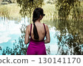 A woman in a magenta jumpsuit enjoying a peaceful moment by a lake. A girl wearing a bright purple pink pants and top gazes at a calm pond back view 130141170