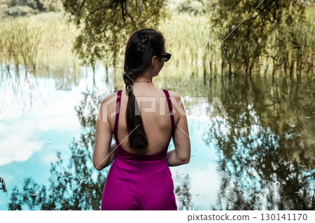 A woman in a magenta jumpsuit enjoying a peaceful moment by a lake. A girl wearing a bright purple pink pants and top gazes at a calm pond back view 130141170