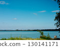 A calm lake reflecting the blue sky and fluffy clouds, surrounded by lush green trees. A serene water in a pond under a blue sky with white clouds. 130141171