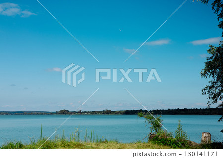 A calm lake reflecting the blue sky and fluffy clouds, surrounded by lush green trees. A serene water in a pond under a blue sky with white clouds. A calm lake reflecting the blue sky and fluffy clouds, surrounded by lush green trees. A serene water in a pond under a blue sky with white clouds. 130141171