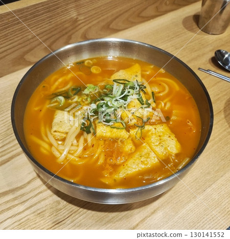 The image shows a bowl of spicy Korean udon noodle soup with fish cakes and green onions. It is a popular comfort food dish. The image shows a bowl of spicy Korean udon noodle soup with fish cakes and green onions. It is a popular comfort food dish. 130141552