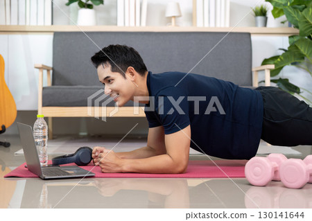 Home Workout. Young man smiling while doing plank exercise with laptop. Home Workout. Young man smiling while doing plank exercise with laptop. 130141644
