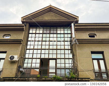 Old building facade with large glass windows framed by geometric grid and metal balcony railings. Architecture, urban design, cultural heritage and city infrastructure. 130142056